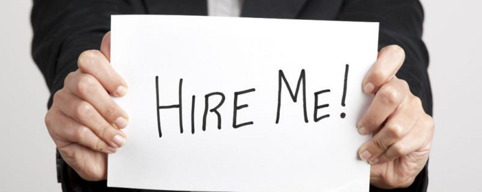 Hands of a person in a blazer and white shirt, holding a white sign that says, "Hire Me!" in black marker.